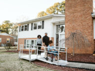 A family of three stands outdoors on a wheelchair ramp leading to the entrance of a house. The person in the wheelchair is being accompanied by two adults.