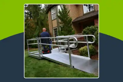 A person in a wheelchair uses a metal accessibility ramp leading to the front door of a house, surrounded by grass and shrubs.