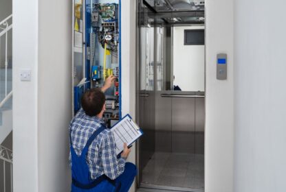 A technician in blue overalls works on the control panel of an elevator, holding a clipboard with a checklist.