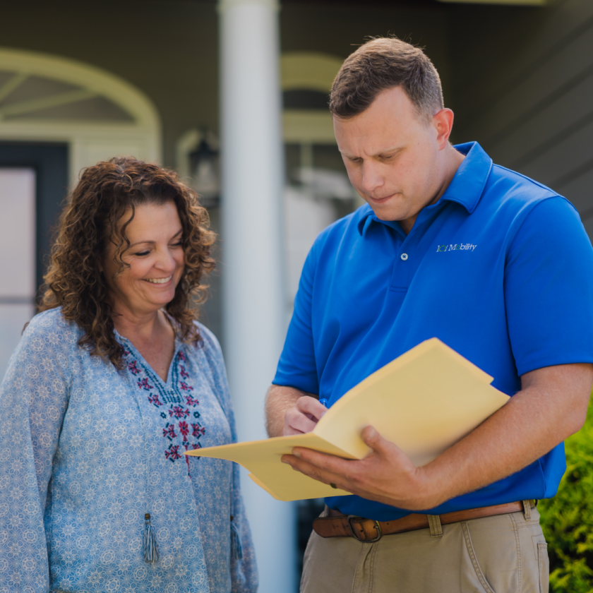 A man in a blue polo shirt shows a document to a woman standing outside a house.