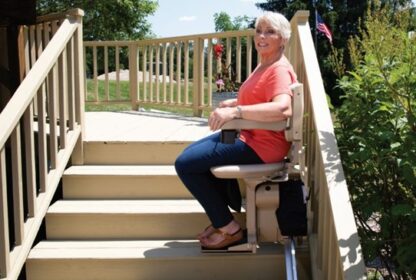 A woman wearing a coral shirt and jeans is sitting on a stairlift chair, ascending an outdoor staircase with wooden railings and greenery in the background.