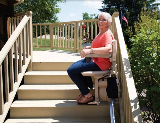 A woman wearing a coral shirt and jeans is sitting on a stairlift chair, ascending an outdoor staircase with wooden railings and greenery in the background.