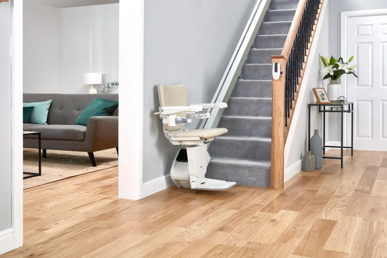 A beige stairlift installed on a carpeted staircase adjacent to a hallway with wooden flooring, leading to a living room with a grey sofa and turquoise pillows.