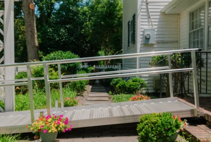 A metal wheelchair ramp leads to a house entrance. The yard has green shrubs, colorful flowers, and a paved walkway.