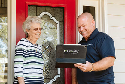 A woman stands at her front door, smiling, while a man in a navy shirt shows her information on a tablet that reads "1CI Mobility.