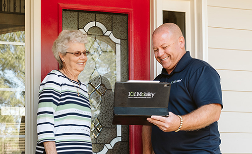 A woman stands at her front door, smiling, while a man in a navy shirt shows her information on a tablet that reads "1CI Mobility.