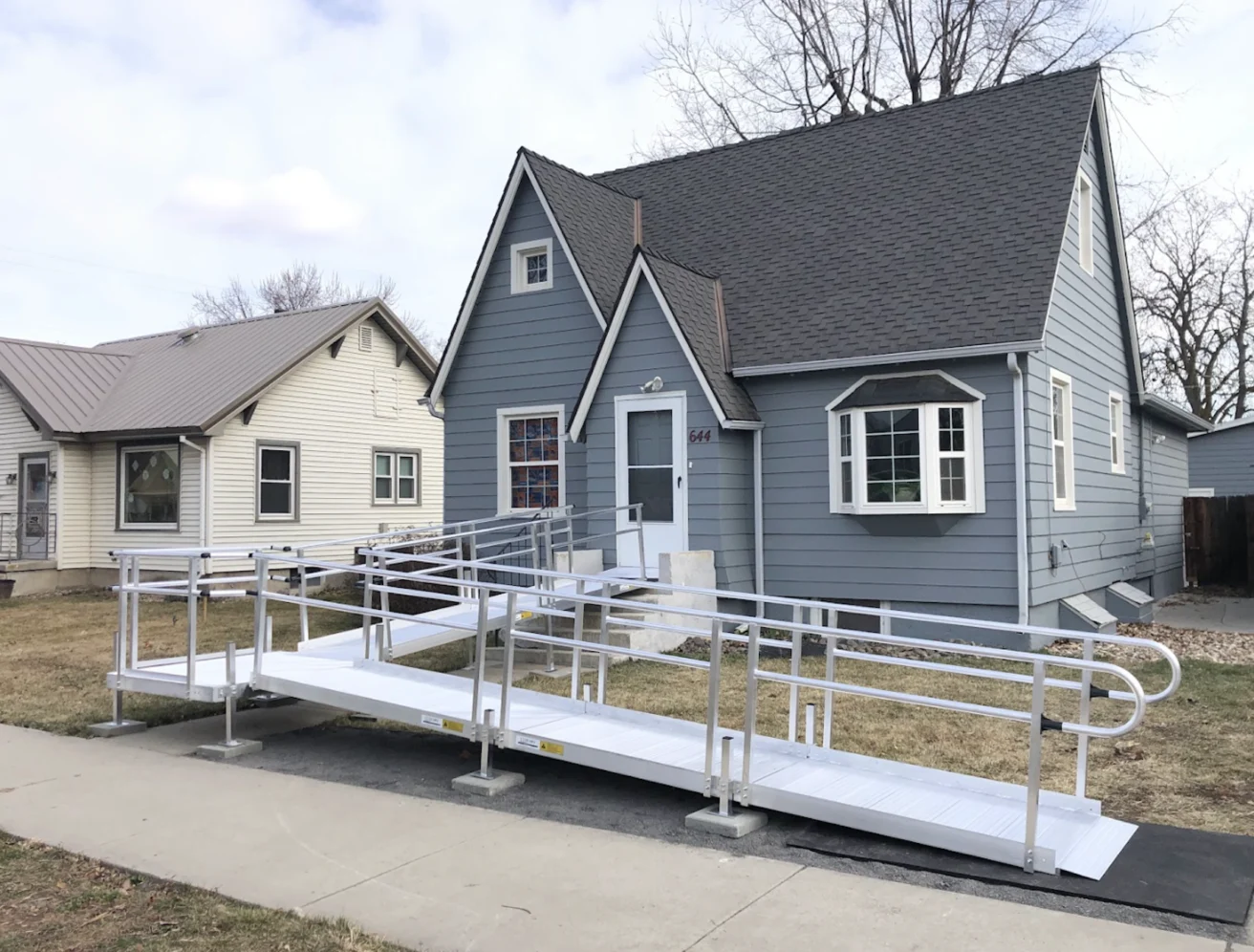 A gray house with white trim has a metal wheelchair ramp leading from the sidewalk to the front door, providing accessible entry.