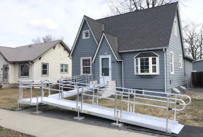 A gray house with white trim has a metal wheelchair ramp leading from the sidewalk to the front door, providing accessible entry.