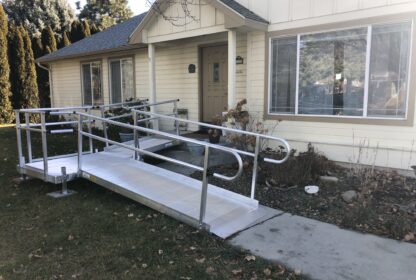 A single-story house with a wide, metal accessibility ramp leading to the front entrance. The ramp passes through a modest front yard with grass and some shrubs.