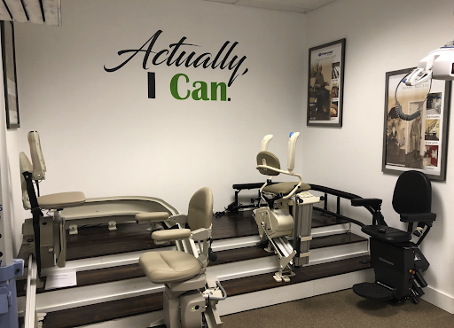 A showroom displays various models of stairlifts on a staircase. A wall sign reads "Actually, I Can." Poster pictures depict stairlift installations in homes.