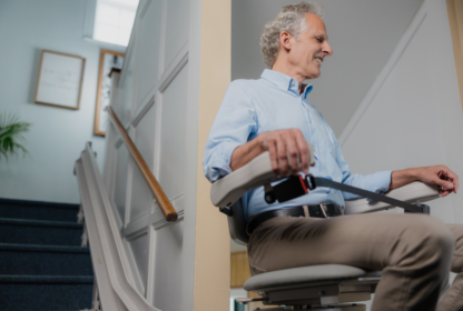 An older man with gray hair and a light blue shirt rides a stairlift chair at home, descending down a staircase.