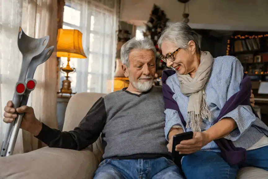Older couple sitting on a couch, with the man holding crutches and the woman showing him something on a smartphone. They appear engaged and content in a cozy living room.