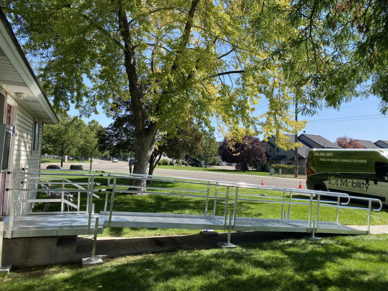 A metal wheelchair ramp attached to a house extends onto a lawn with a tree. In the background, there are residential buildings and a vehicle with "Mobility" written on its side.