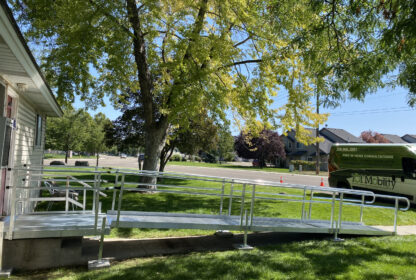 A metal wheelchair ramp attached to a house extends onto a lawn with a tree. In the background, there are residential buildings and a vehicle with "Mobility" written on its side.