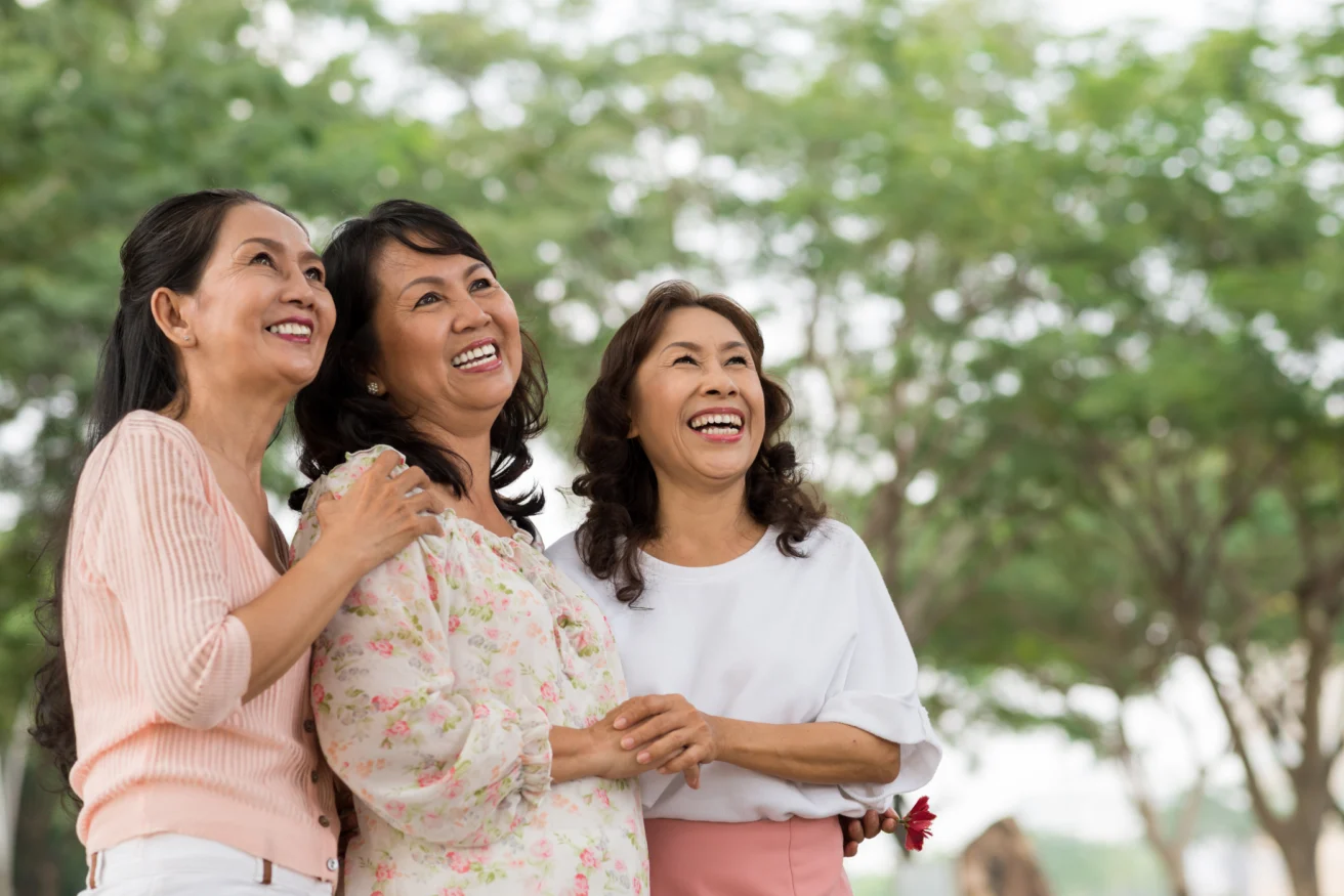 Three women stand outdoors, smiling and laughing, with trees in the background.