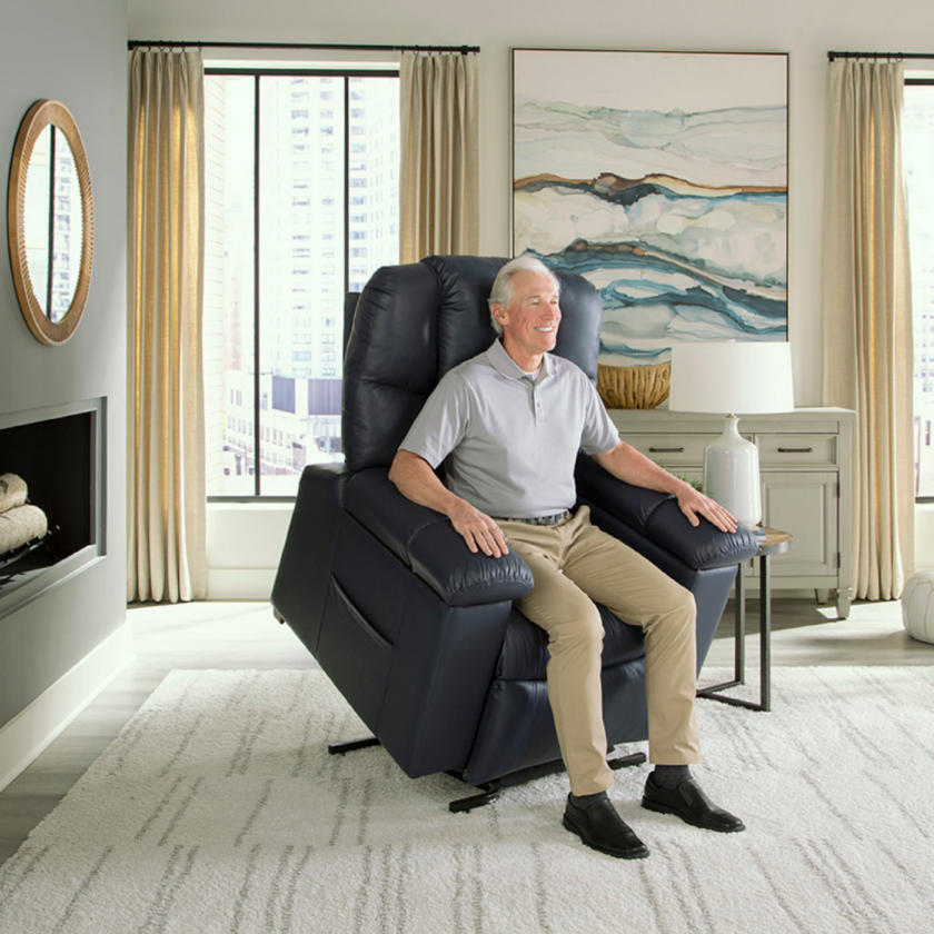An elderly man sits in a blue recliner chair in a brightly lit living room with large windows, beige curtains, a wall mirror, and modern decor.