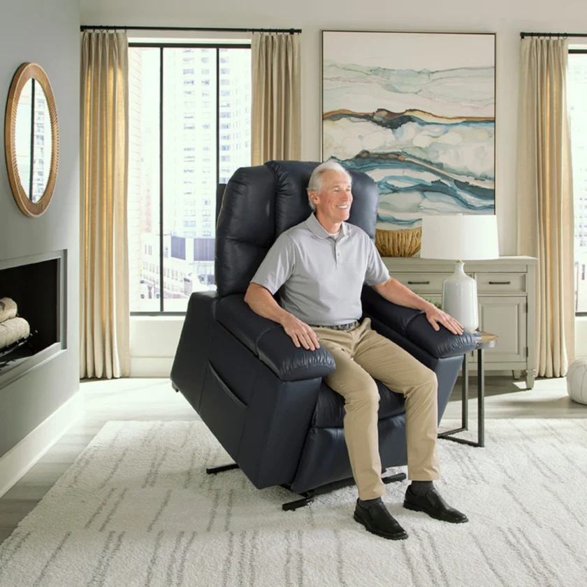 An elderly man sits in a blue recliner chair in a brightly lit living room with large windows, beige curtains, a wall mirror, and modern decor.