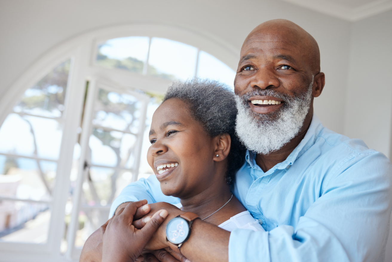Two people smiling and embracing in a bright room with arched windows. The person in front has short gray hair, and the person behind has a gray beard and wears a light blue shirt.