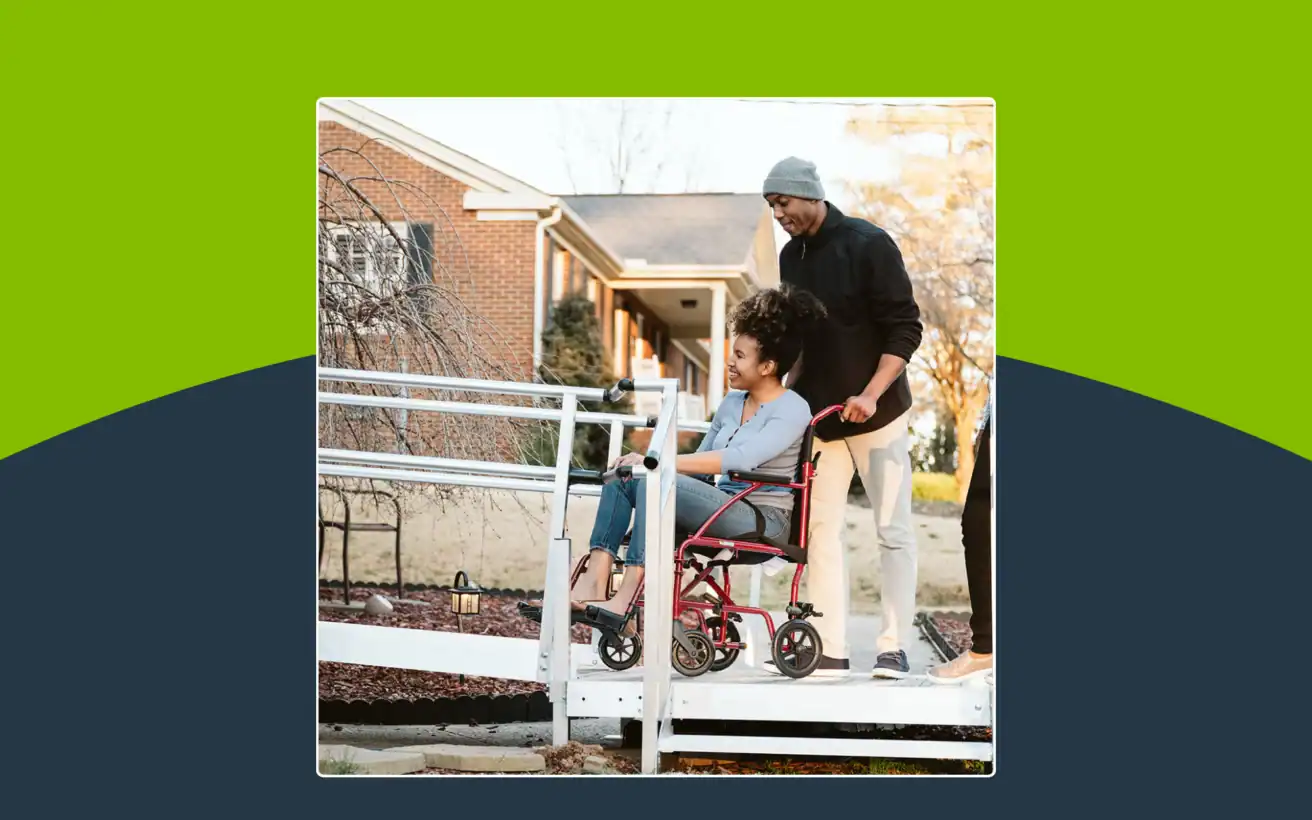 A person in a wheelchair is being assisted by another person up a ramp outside a brick house on a sunny day.