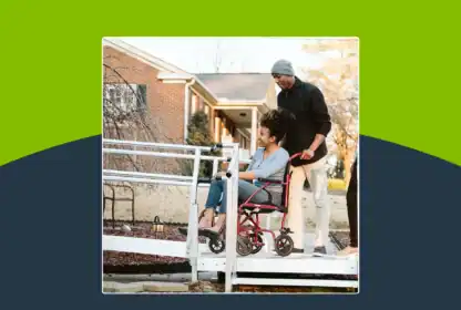 A person in a wheelchair is being assisted by another person up a ramp outside a brick house on a sunny day.