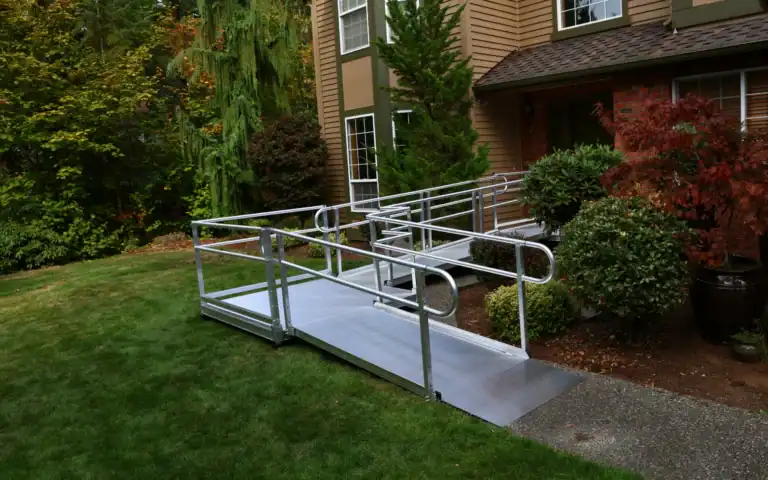 A metal wheelchair ramp with railings leads from a sidewalk to the front door of a house, surrounded by grass and landscaped bushes.