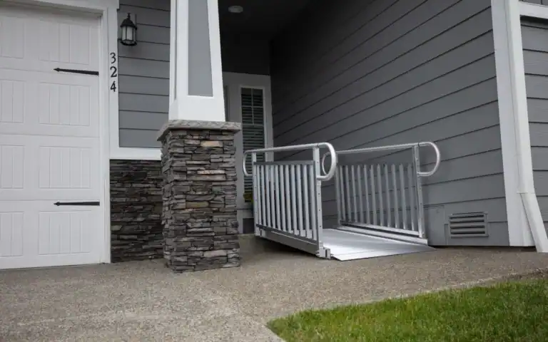 A portable wheelchair ramp leads from the sidewalk up to the entrance of a grey house with stone accent and white door.