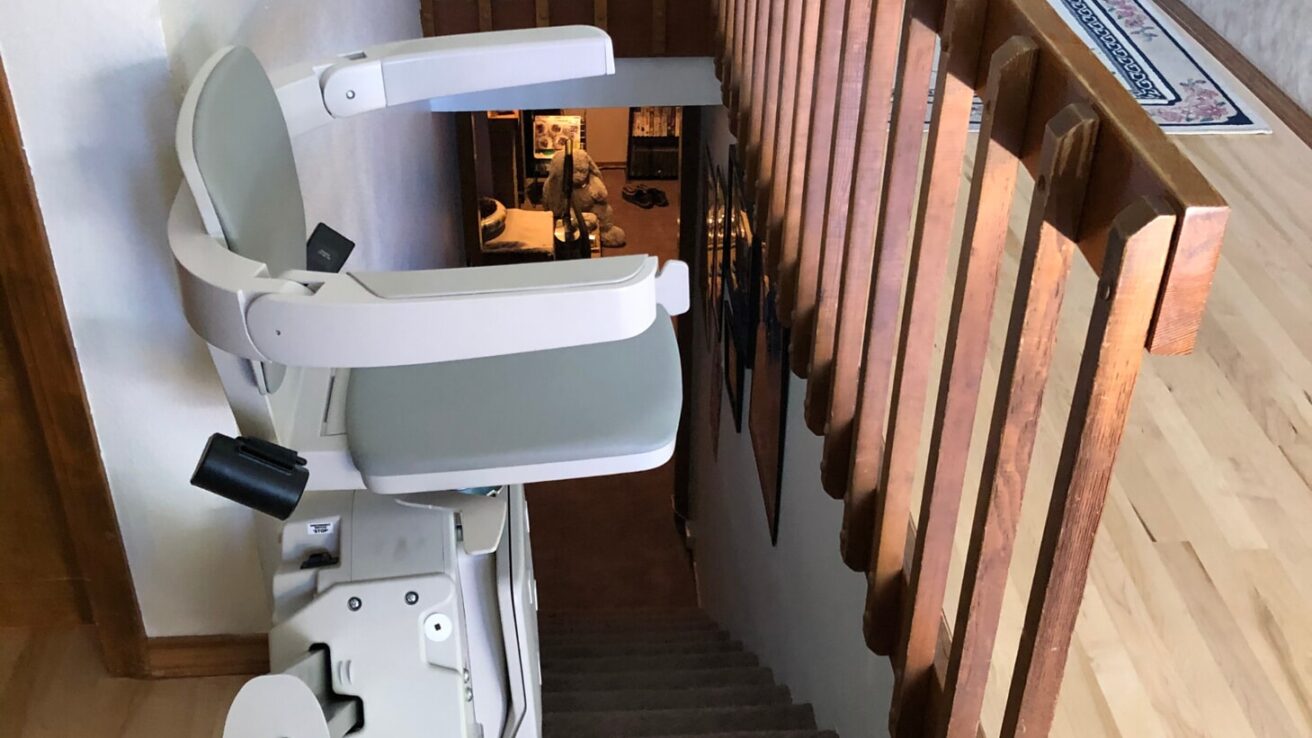 A chair lift is installed along the staircase of a home, providing accessibility between floors. The stair lift has a grey and white seat and runs parallel to the wooden handrail.