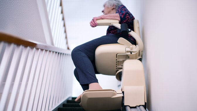 An elderly person sits on a stairlift, ascending a narrow staircase, with one hand resting on the armrest.