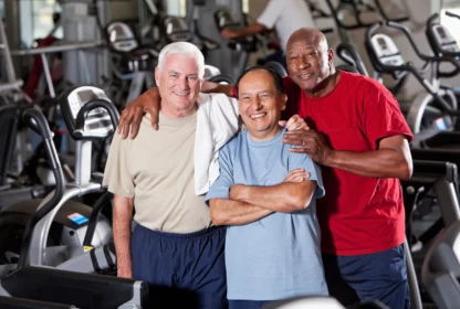 Three men standing together in a gym, smiling, with exercise equipment in the background. One man has a towel draped over his shoulder.