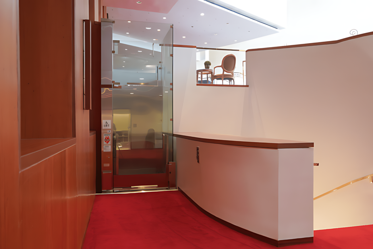 A modern interior with a red carpeted floor features a stair lift and a glass door on the left, leading to an upper floor with a wooden railing and chairs visible in the background.