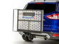 A metal wheelchair carrier with a silver star pattern is attached to the back of a blue SUV. The carrier displays a "Harmar" logo and phone number.