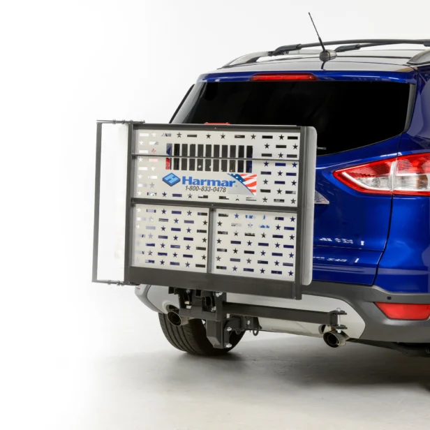 A metal wheelchair carrier with a silver star pattern is attached to the back of a blue SUV. The carrier displays a "Harmar" logo and phone number.