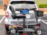 A mobility scooter is mounted on a lift attached to the back of a silver SUV parked in a lot.