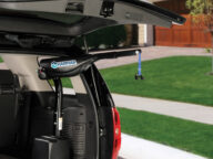 An SUV with the trunk open, showing a Harmar lift installed inside, designed to assist in lifting a power wheelchair or scooter. The vehicle is parked in a residential driveway.