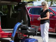 An elderly woman operates a mechanical lift to load a mobility scooter into the back of a red minivan in a parking lot.