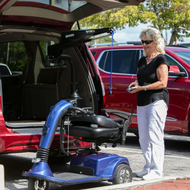 An elderly woman operates a mechanical lift to load a mobility scooter into the back of a red minivan in a parking lot.