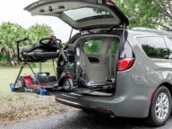 A mobility scooter is secured on a lift attached to the back of an open gray minivan. The lift is in the process of lowering the scooter to the ground. Trees and grass are visible in the background.