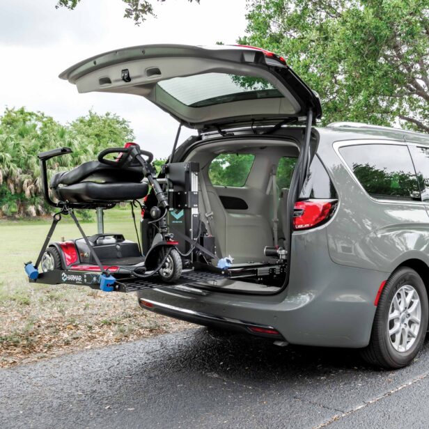 A mobility scooter is secured on a lift attached to the back of an open gray minivan. The lift is in the process of lowering the scooter to the ground. Trees and grass are visible in the background.