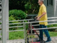 An older man wearing a yellow shirt and blue jeans uses a walker to navigate a wheelchair ramp outside a house with a garden.