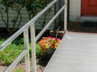 A metal accessibility ramp leads to a house with an orange door. Flowers and greenery are visible beside the ramp.