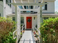 A white house with a red door and a wreath. A ramp leads to the entrance under a white pergola labeled "6424." Green bushes line the pathway.