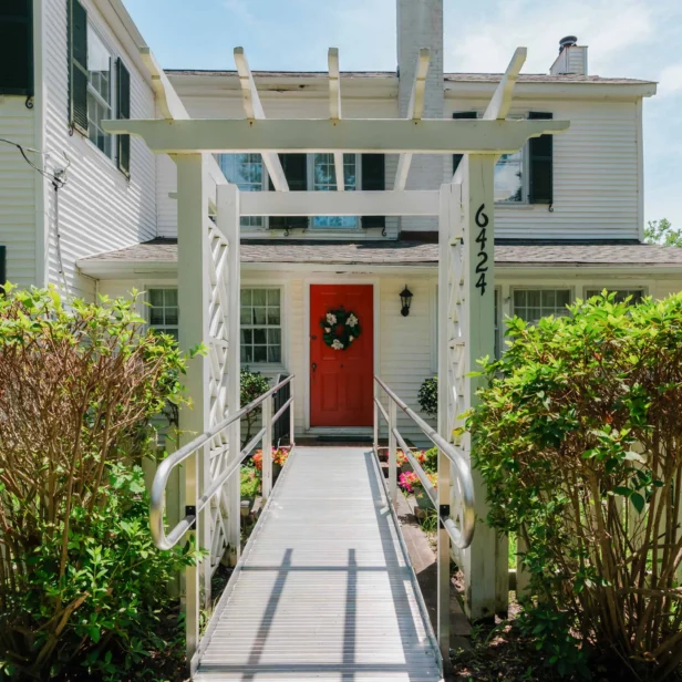 A white house with a red door and a wreath. A ramp leads to the entrance under a white pergola labeled "6424." Green bushes line the pathway.