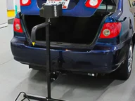 A blue compact car with an open trunk is parked in a garage. A metal device is positioned behind the vehicle, likely for testing or support purposes.