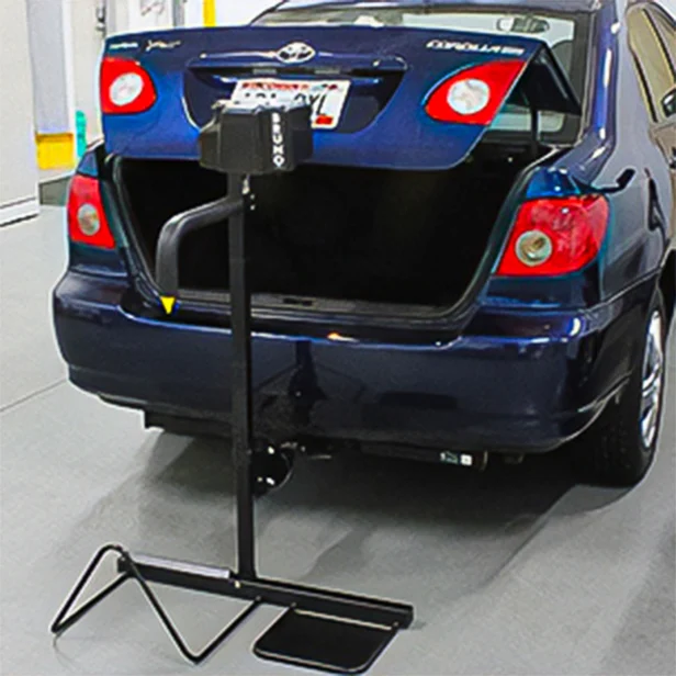 A blue compact car with an open trunk is parked in a garage. A metal device is positioned behind the vehicle, likely for testing or support purposes.