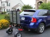 A blue SUV is parked in a residential driveway with a mobility scooter lift attached to the rear. The lift is in an upright, folded position. Houses and a garden are visible in the background.