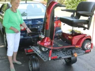 An elderly woman stands next to a blue car, securing a red mobility scooter onto a rear platform carrier in a driveway.
