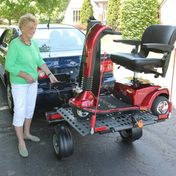 An elderly woman stands next to a blue car, securing a red mobility scooter onto a rear platform carrier in a driveway.