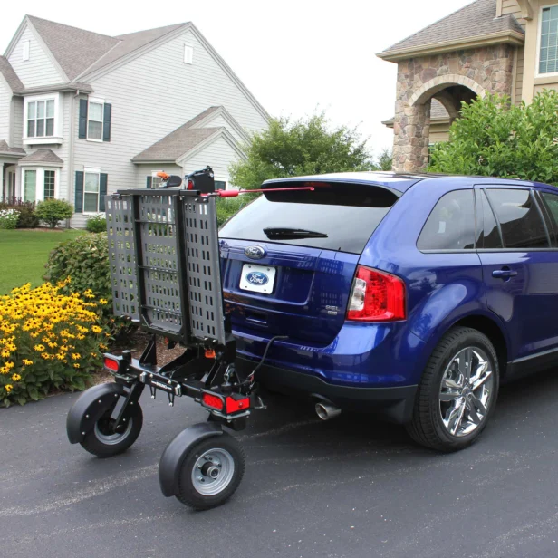 A blue SUV is parked in a residential driveway with a mobility scooter lift attached to the rear. The lift is in an upright, folded position. Houses and a garden are visible in the background.