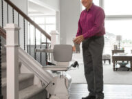 A smiling older man in a magenta shirt and gray pants is standing next to a stairlift at the bottom of a staircase in a well-lit home.