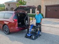 A person stands beside a parked red minivan, operating a lift to load a blue mobility scooter into the vehicle's open trunk. The scene is set in a residential driveway in front of a large house.